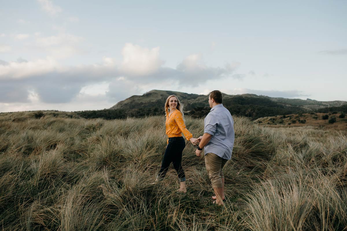 couple walking along muriwai beach auckland new zealand during a golden light evening lifestyle engagment pre-wedding photoshoot session by sarah weber photography