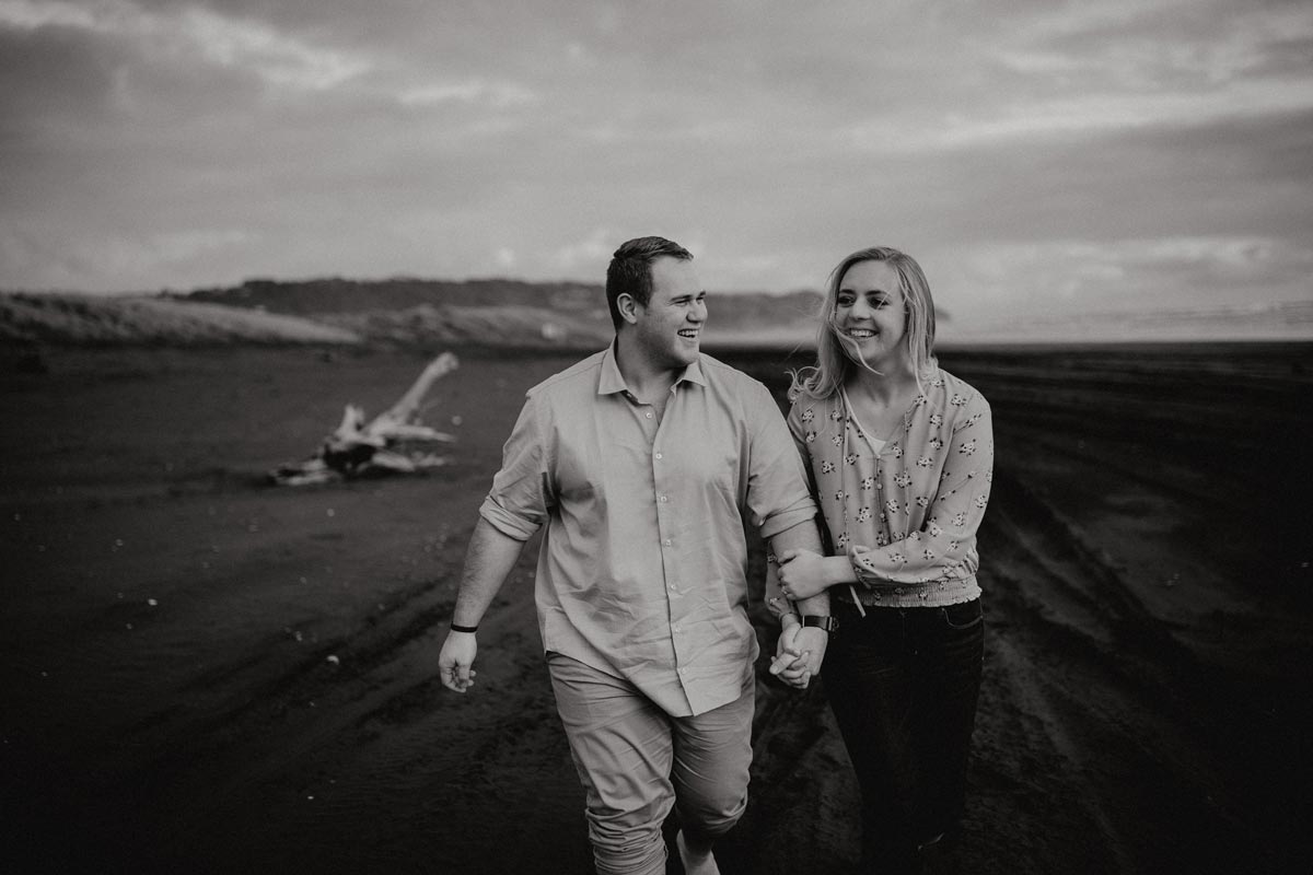 happy couple walking along muriwai beach auckland new zealand during a golden light evening lifestyle engagment pre-wedding photoshoot session with sarah weber photography