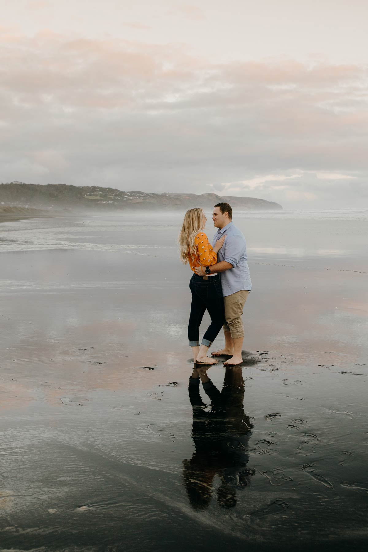 couple embracing with a wet sand reflection during a golden light evening lifestyle engagment pre-wedding photoshoot session at muriwai beach auckland new zealand photos by sarah weber photography